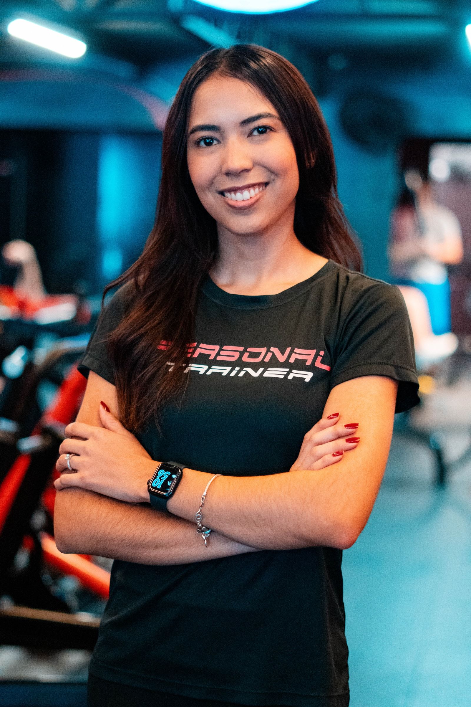 Camila Tenório sorrindo, de braços cruzados, com camiseta escrita Personal Trainer, em uma academia.